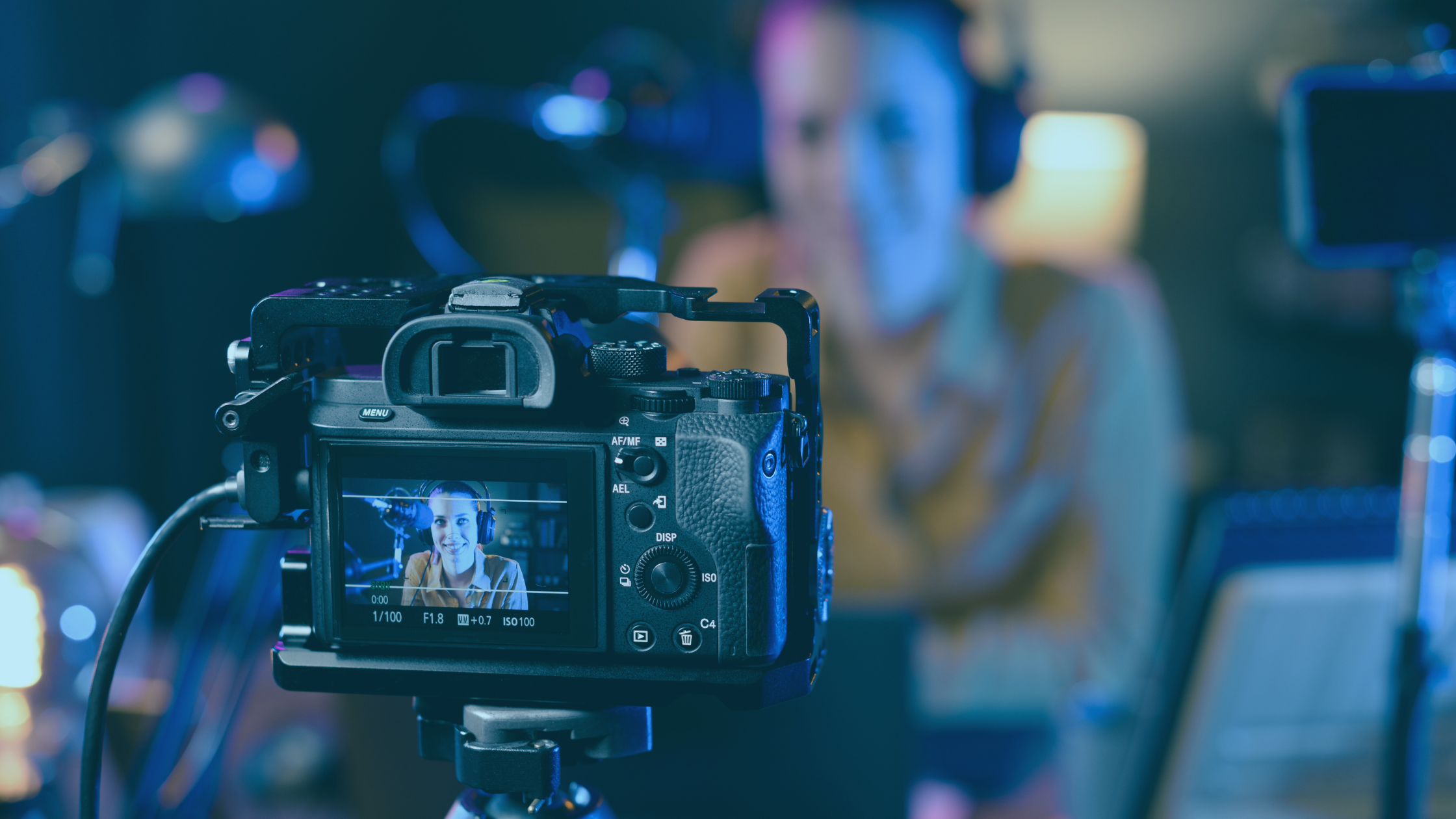 Em foco temos uma câmera, mais ao fundo, desfocada, há uma pessoa gravando um vídeo.